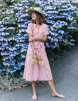A woman holds her sandals while standing barefoot nest to a flowering bush